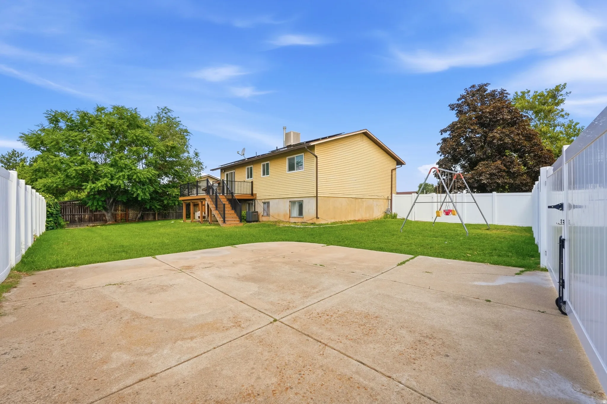 Spacious backyard featuring a large concrete patio, manicured lawn, white vinyl fencing, mature trees, and a multi-level wood deck with stairs