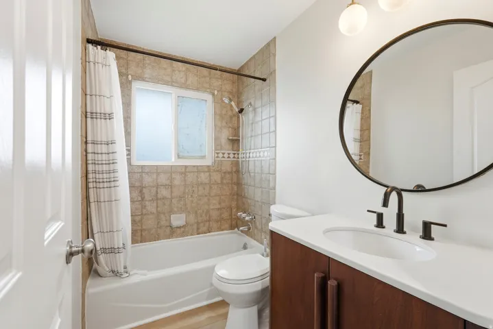 Bathroom featuring a modern vanity with integrated sink, a large round mirror, dark bronze fixtures, and a tiled shower-tub combination with an accent border