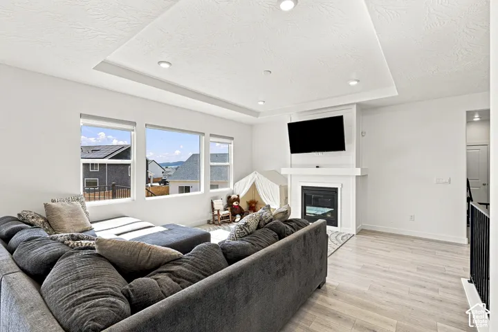 Spacious living area featuring a tray ceiling with recessed lighting, large windows, a fireplace with a white mantel, and light wood-finish flooring