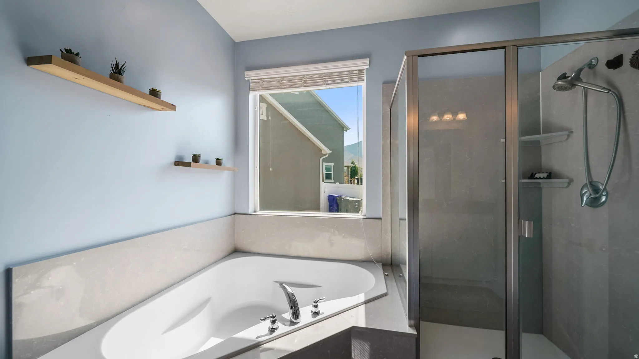 Soaking tub with integrated surround, framed window with blinds, glass-enclosed shower with tiled walls, and wall-mounted wooden shelving