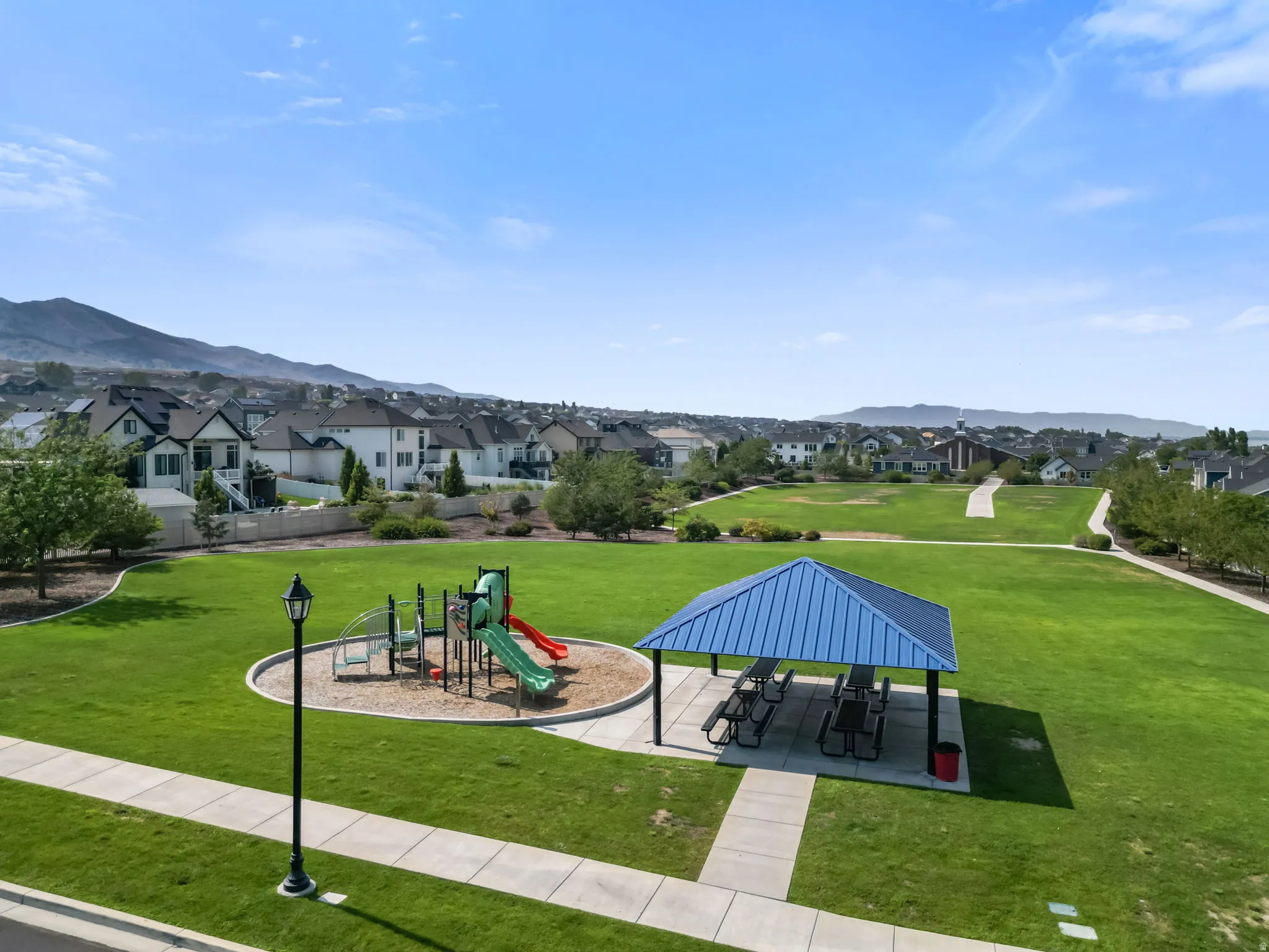 Expansive green space featuring a playground structure, covered picnic area with tables, and paved walking paths
