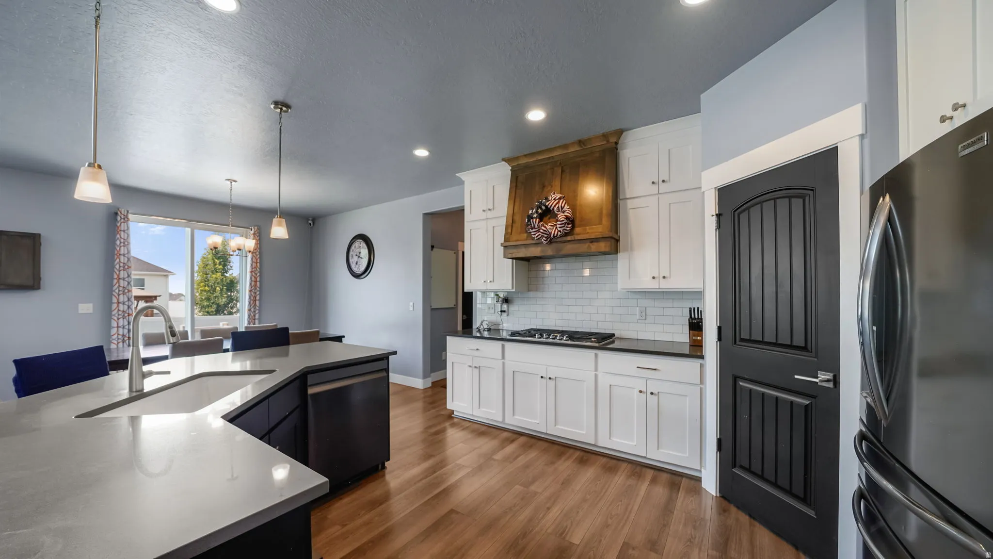 Kitchen featuring a wood-finish range hood, white subway tile backsplash, white shaker cabinetry, stainless steel appliances, and a kitchen island with a built-in sink