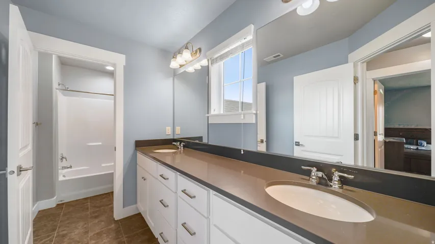 Dual sink vanity with white cabinetry, brown tile flooring, and a white tub with shower