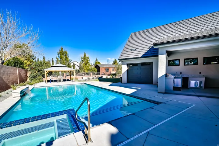 Outdoor living space featuring a swimming pool with integrated spa and waterfall feature, expansive concrete patio, built-in outdoor kitchen with sink and refrigeration, and a shaded gazebo structure