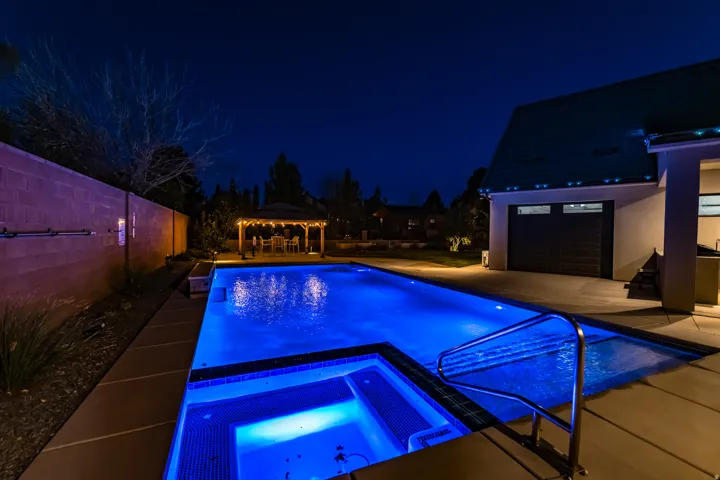 Illuminated backyard featuring an in-ground swimming pool and integrated spa, a wooden gazebo, concrete patio, and block wall fencing