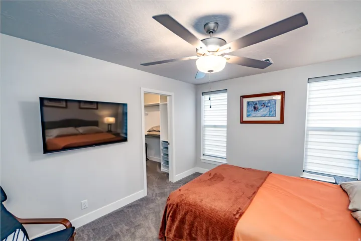 Bedroom featuring a mounted television, a ceiling fan with integrated lighting, two windows with blinds, and carpeted flooring