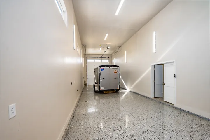 Spacious garage featuring a roll-up garage door, epoxy-coated flooring, multiple fluorescent light fixtures, upper transom windows, and a doorway with a white paneled door