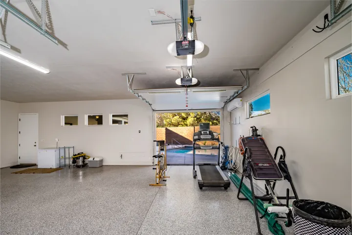 Spacious garage featuring a durable epoxy flooring system, multiple windows for natural light, a white side-entry door, and an open garage door revealing an outdoor pool