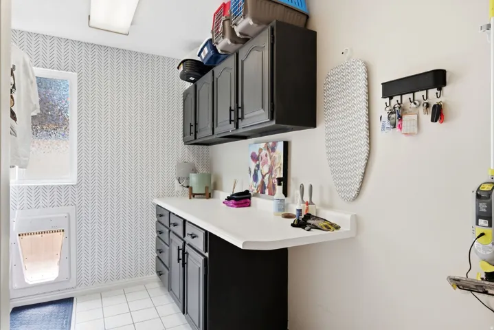 Utility room featuring patterned wallpaper, white tile flooring, dark cabinetry, and white countertops
