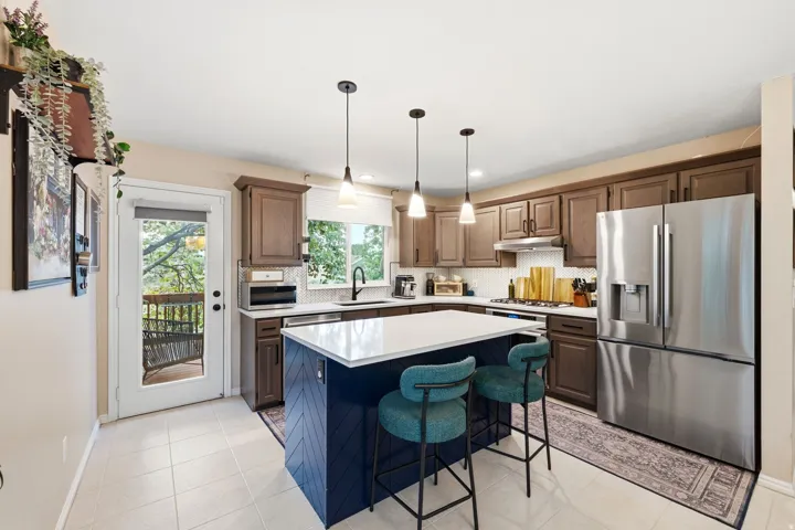 Kitchen featuring a blue-paneled island with a white countertop, stainless steel appliances, wood-finish cabinetry, tile flooring, and a glass-paneled door leading to an outdoor deck