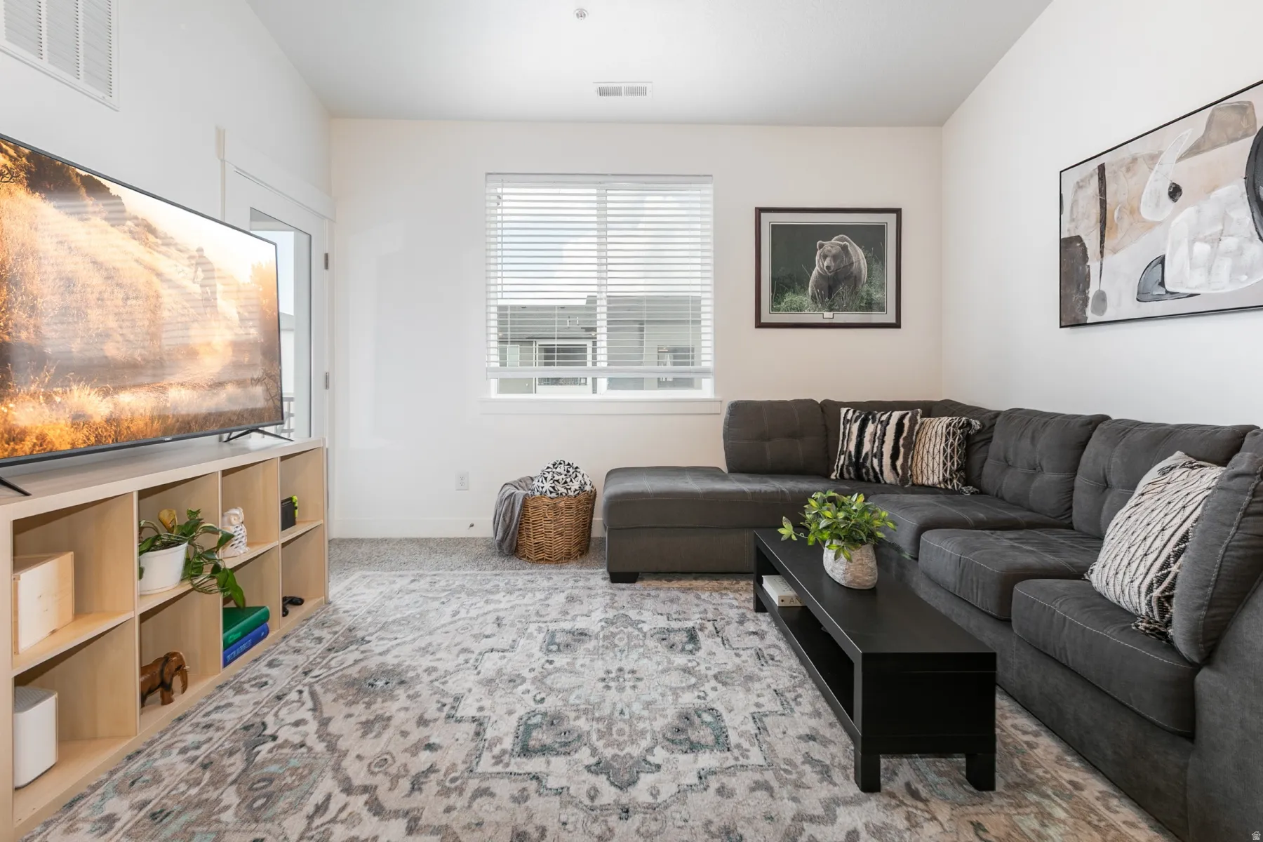 Bright living area featuring light-colored walls, carpeted flooring, and a large window with horizontal blinds