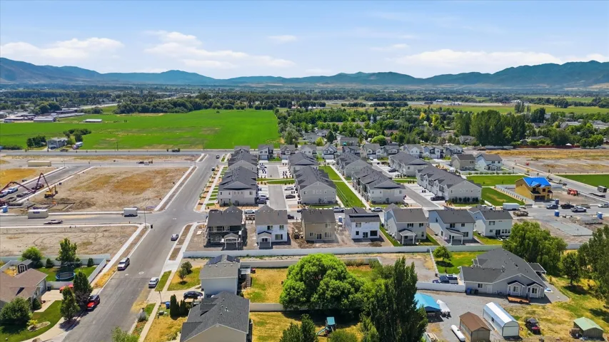 Expansive residential development featuring newly constructed homes with dark roofs, surrounded by open green fields and distant mountains