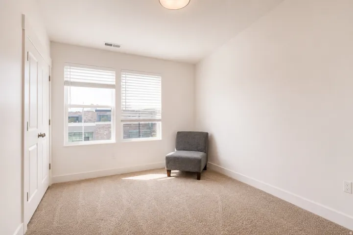 Plush carpet flooring, light-colored walls, two large windows with blinds, and a paneled door with dark hardware