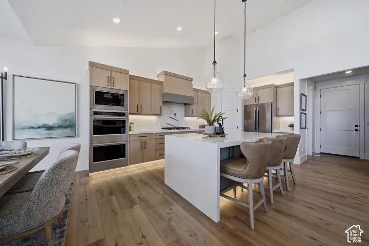 Modern kitchen featuring a large waterfall island, stainless steel built-in appliances, light wood-finish cabinetry, and wood-finish flooring