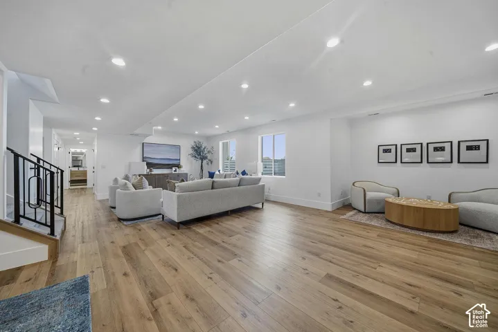 Expansive living area with wood-finish flooring, recessed lighting, white walls, multiple windows, and a staircase with a black metal railing
