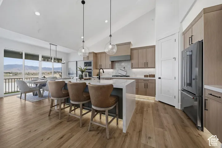 Open-concept kitchen and dining area featuring natural wood cabinetry, stainless steel appliances, a large waterfall-edge island with seating for multiple guests, pendant lighting, wide-plank wood-finish flooring, and expansive windows overlooking mountai