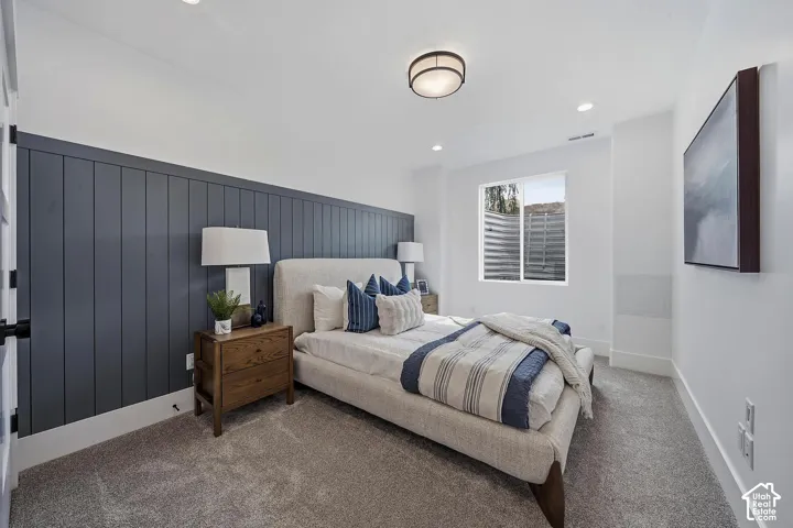 Spacious bedroom featuring a charcoal vertical shiplap accent wall, neutral upholstered bed frame, wood-finish nightstand with table lamp, carpeted flooring, recessed ceiling lighting, and dual windows with plantation shutters allowing natural light throu