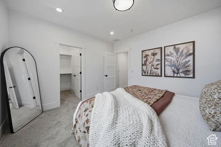 Spacious bedroom featuring neutral carpeting, a bed with coordinating linens and throw blanket, dual framed botanical artwork, an arched mirror, and access to an ensuite bathroom and walk-in closet