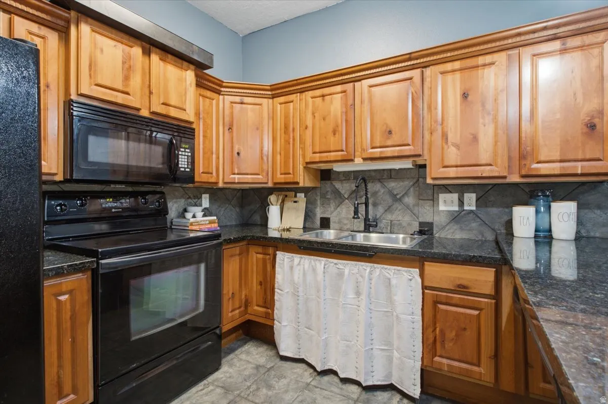 U-shaped kitchen featuring wood cabinetry, black appliances, dark countertops, a stainless steel double sink with a matte black faucet, and a tiled backsplash