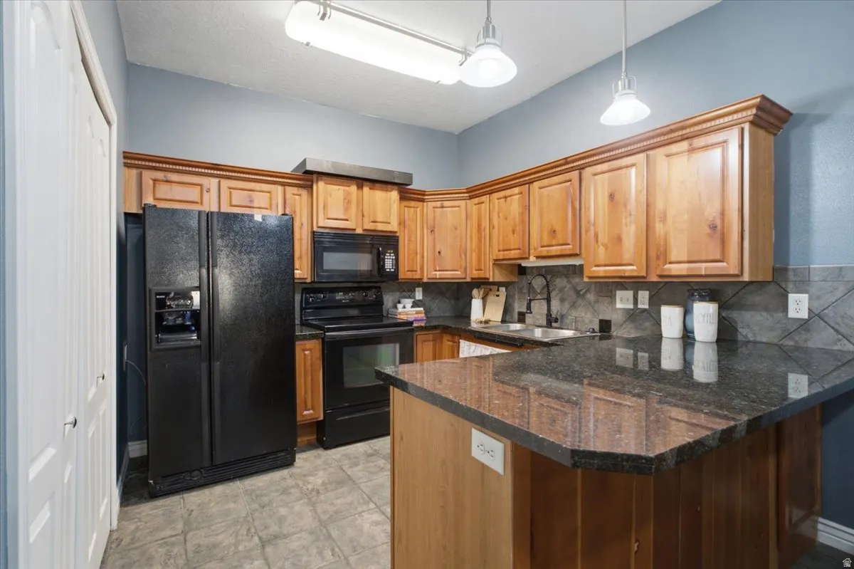 Kitchen featuring wood cabinetry, black appliances, dark countertops, a tile backsplash, and overhead pendant lighting