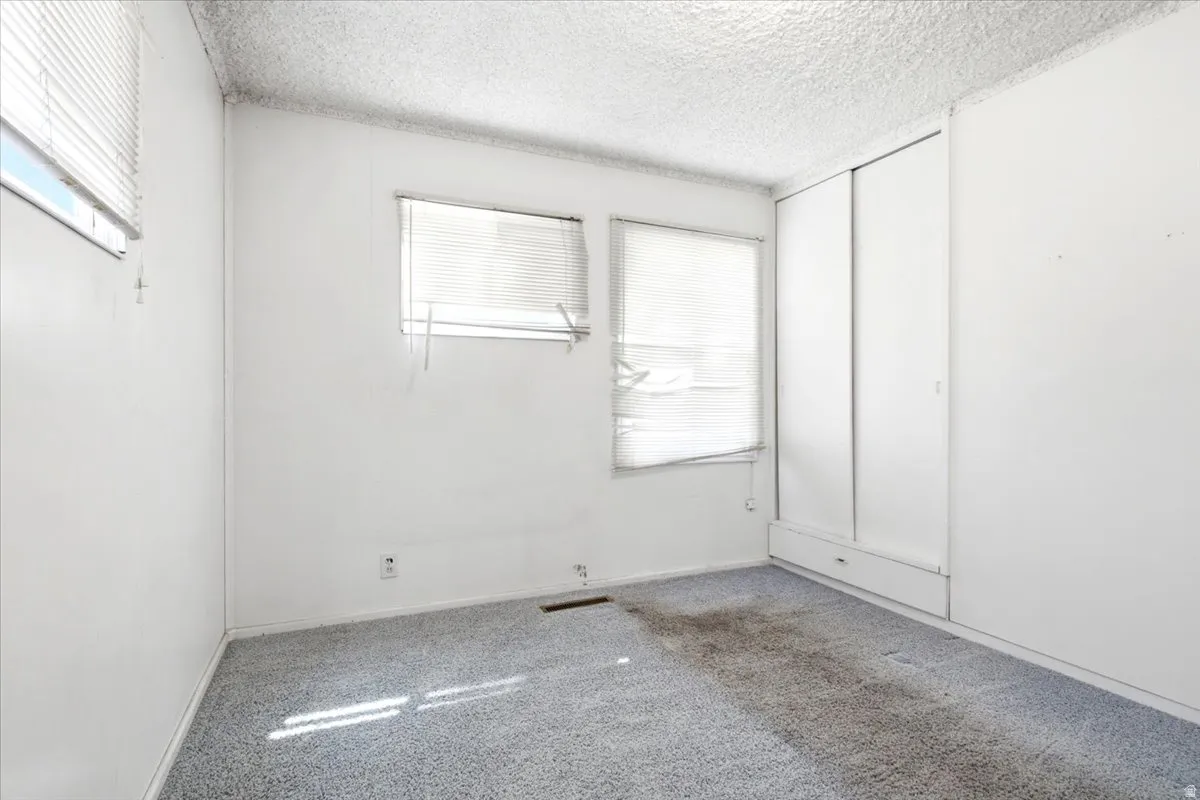 Room featuring two windows with horizontal blinds, built-in sliding wardrobe with lower drawers, textured white ceiling, and light blue carpet flooring