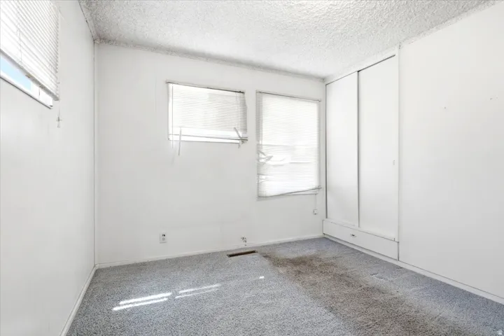 Room featuring two windows with horizontal blinds, built-in sliding wardrobe with lower drawers, textured white ceiling, and light blue carpet flooring