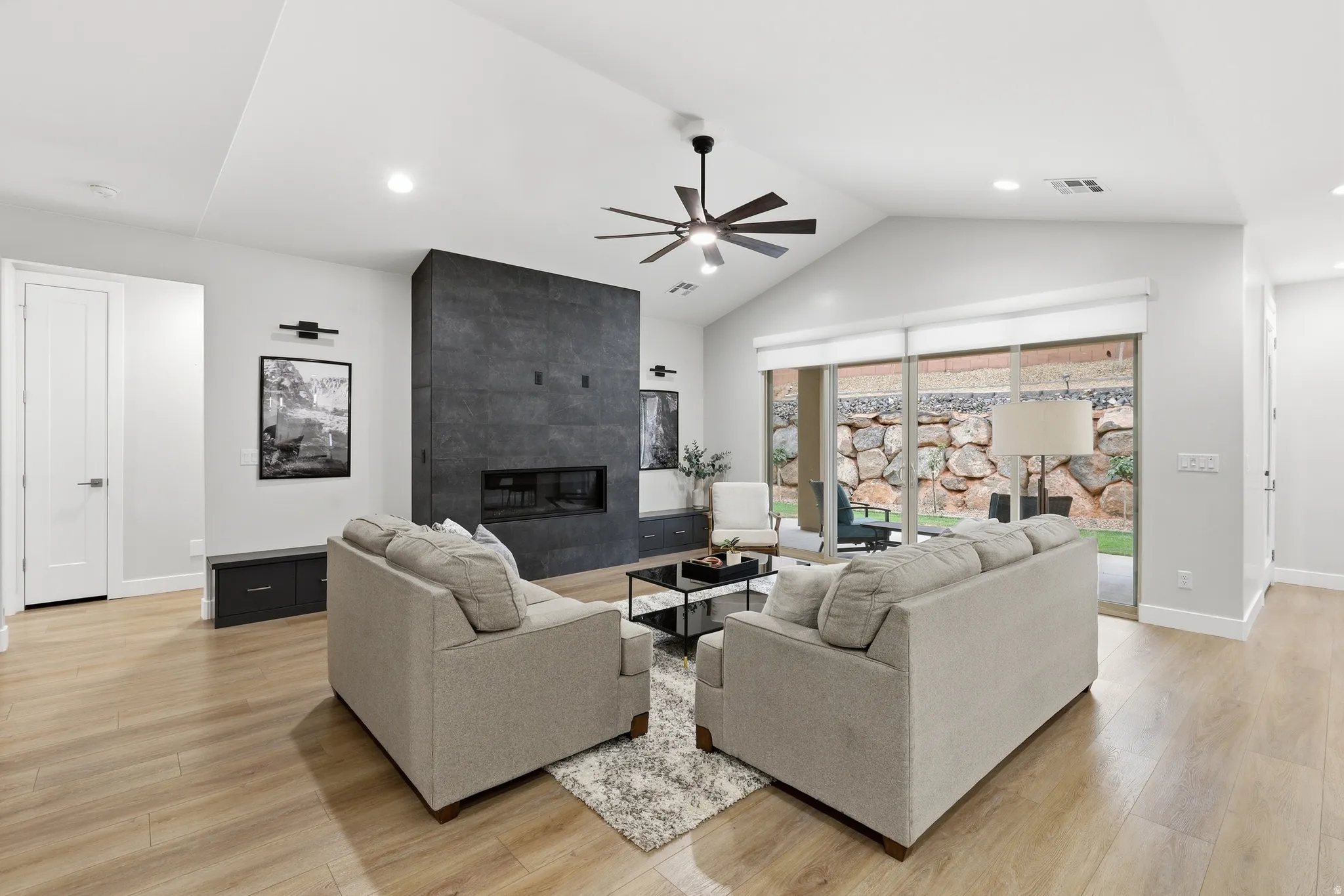 Spacious living area featuring vaulted ceilings, wood-finish flooring, a contemporary fireplace with dark stone surround, a ceiling fan, and sliding glass doors opening to a hardscaped outdoor area