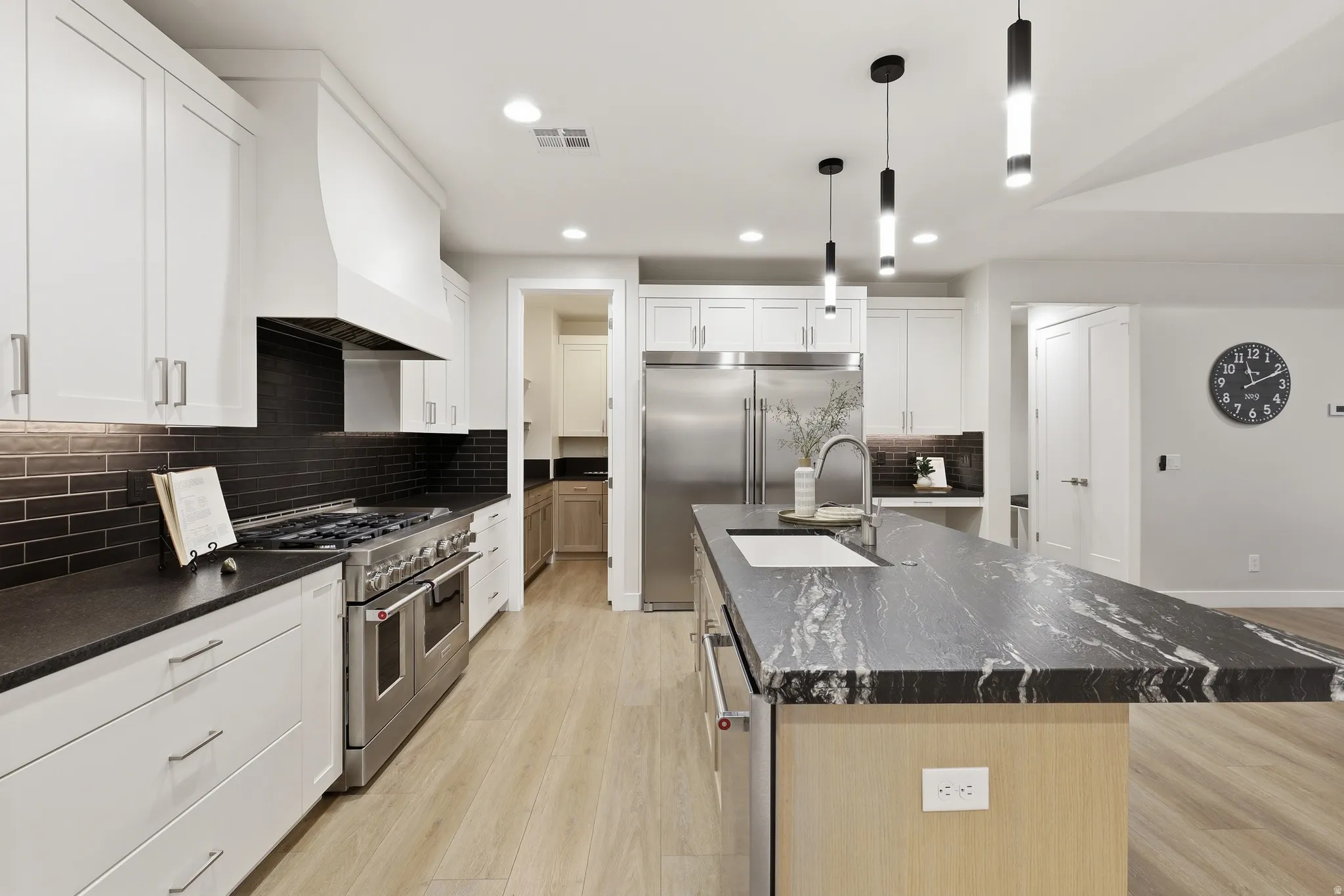 Modern kitchen featuring a large island with a patterned countertop, a stainless steel range with a custom hood, light wood-finish flooring, white upper and lower cabinetry, and a dark subway tile backsplash