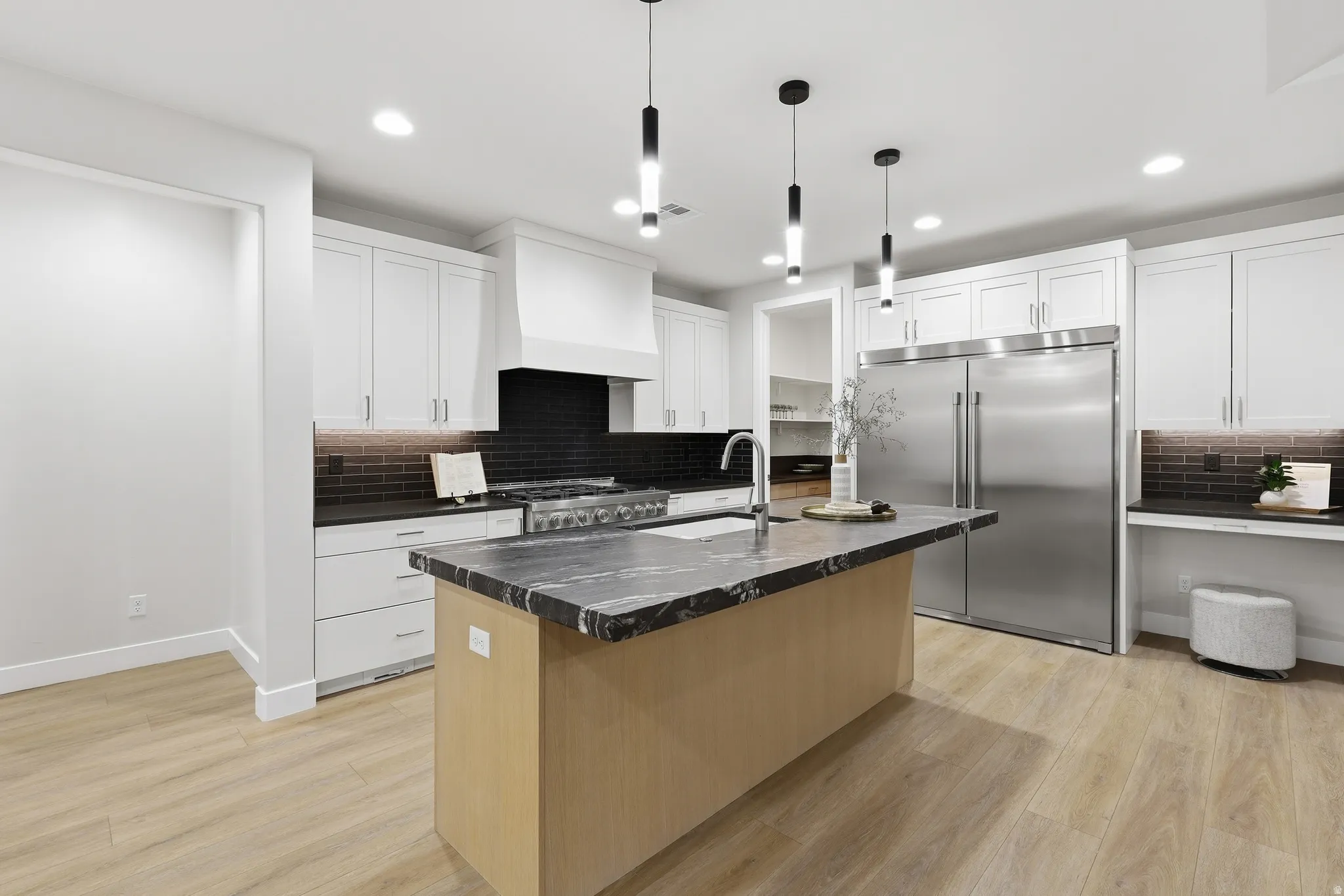Modern kitchen featuring a large central island with a contrasting dark countertop, light wood-finish flooring, white upper and lower cabinetry, stainless steel appliances, and a dark tile backsplash