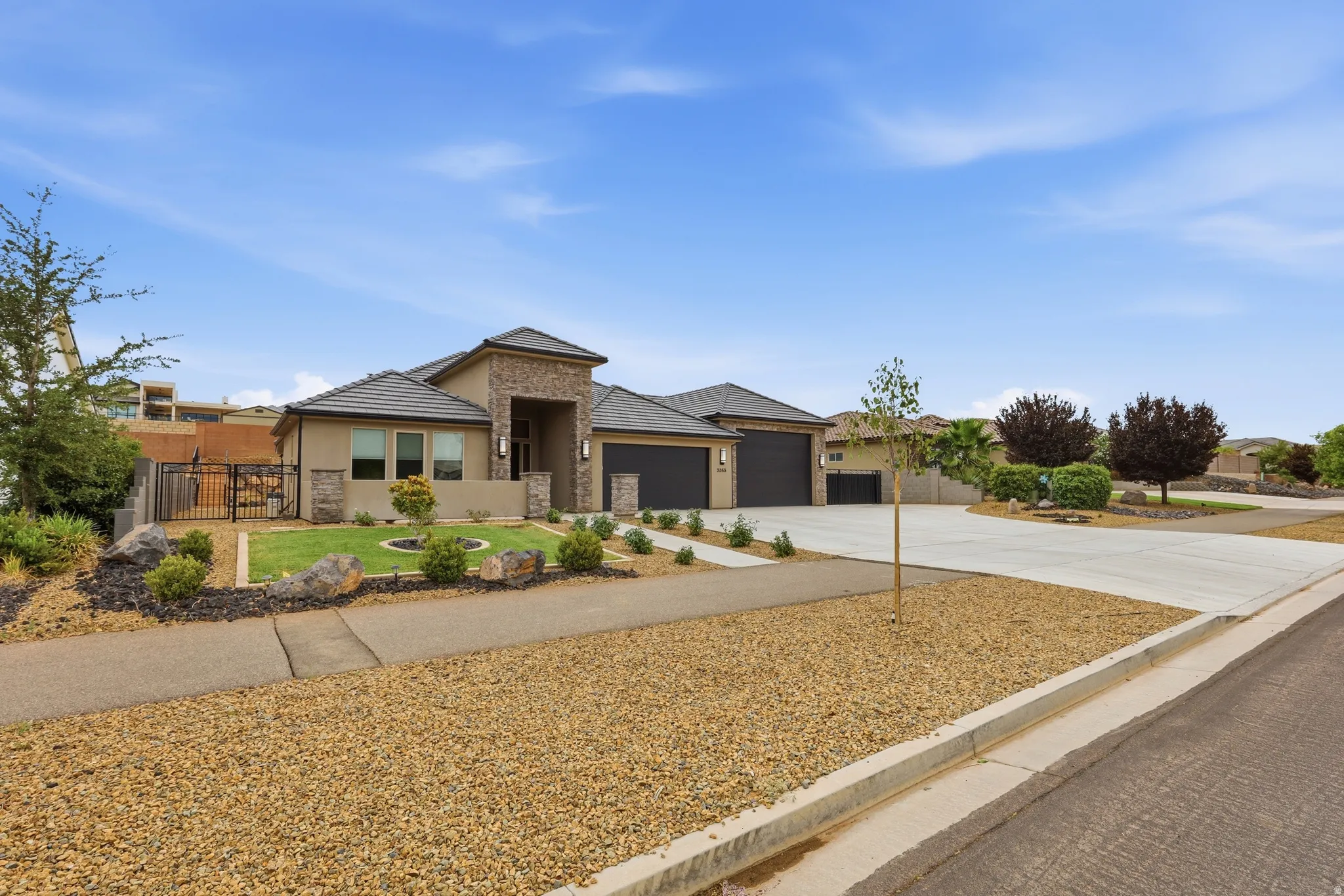 Contemporary stucco exterior featuring stone accents, a multi-car driveway, dark garage doors, a manicured front lawn, and mature landscaping