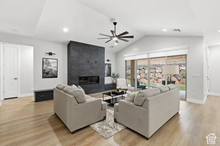 Spacious living area featuring vaulted ceilings, wood-finish flooring, a contemporary fireplace with dark stone surround, a ceiling fan, and sliding glass doors opening to a hardscaped outdoor area