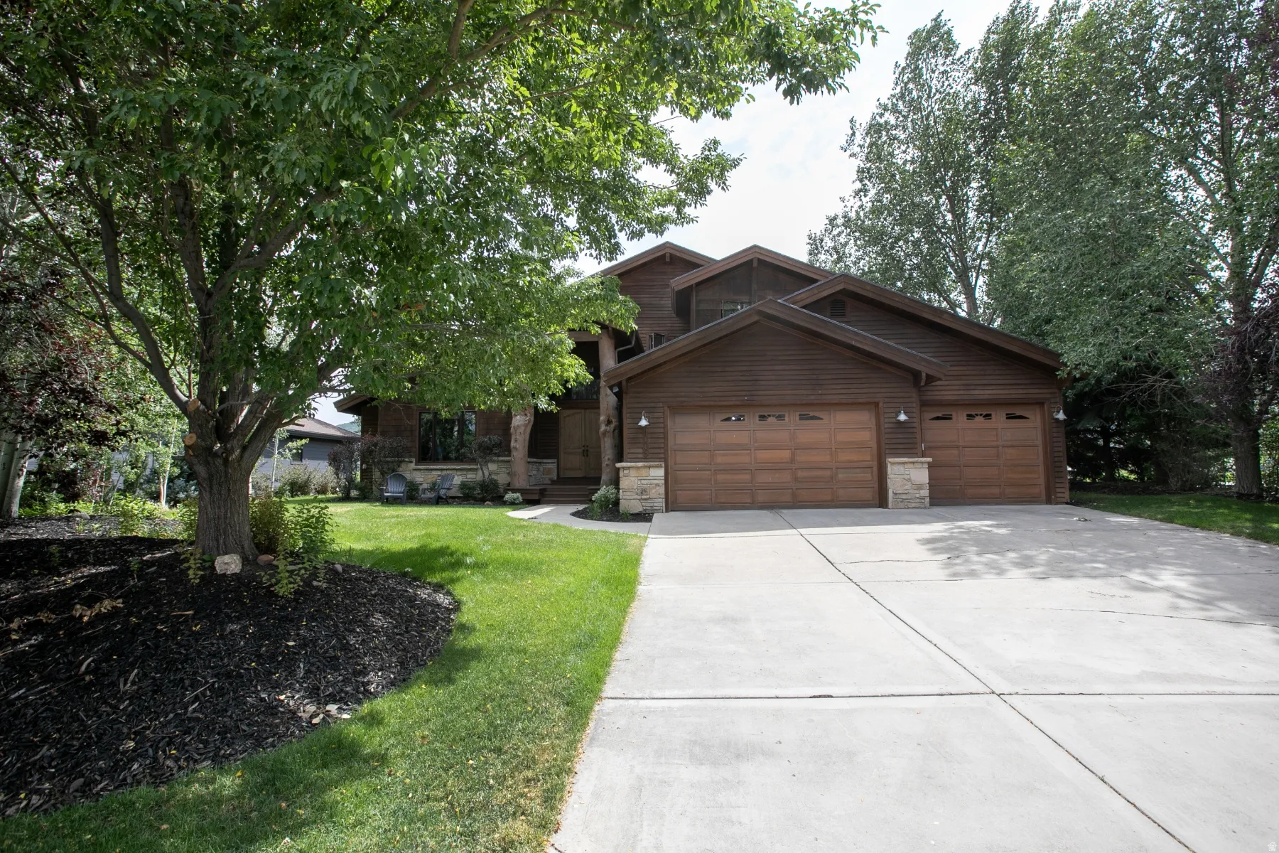 Exterior featuring wood-finish siding, a two-car garage with wood-paneled doors, stone accents, a concrete driveway, and mature landscaping