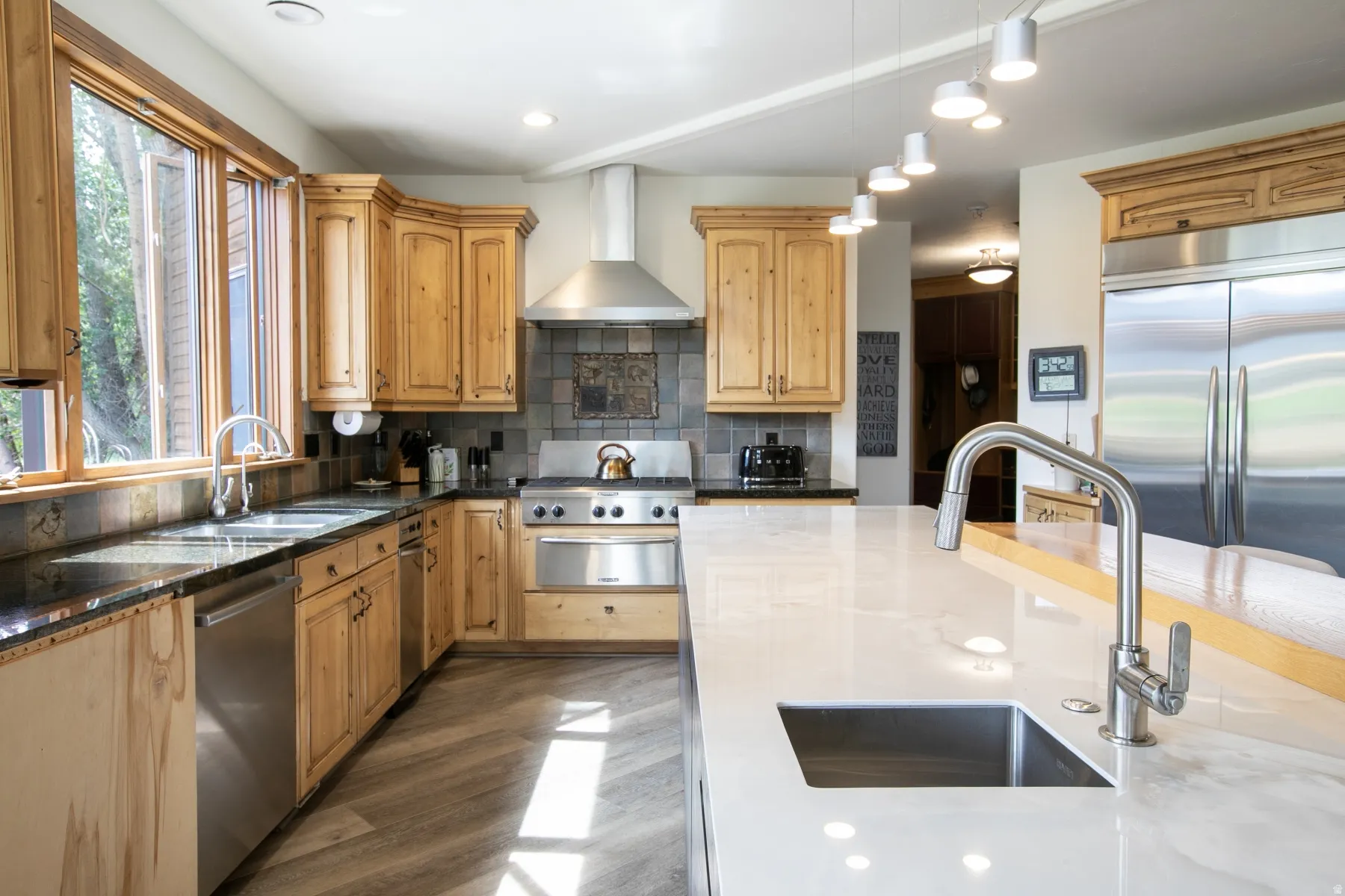 Kitchen featuring light wood cabinetry, stainless steel appliances, a large center island with an integrated sink, dark countertops, and wood-finish flooring