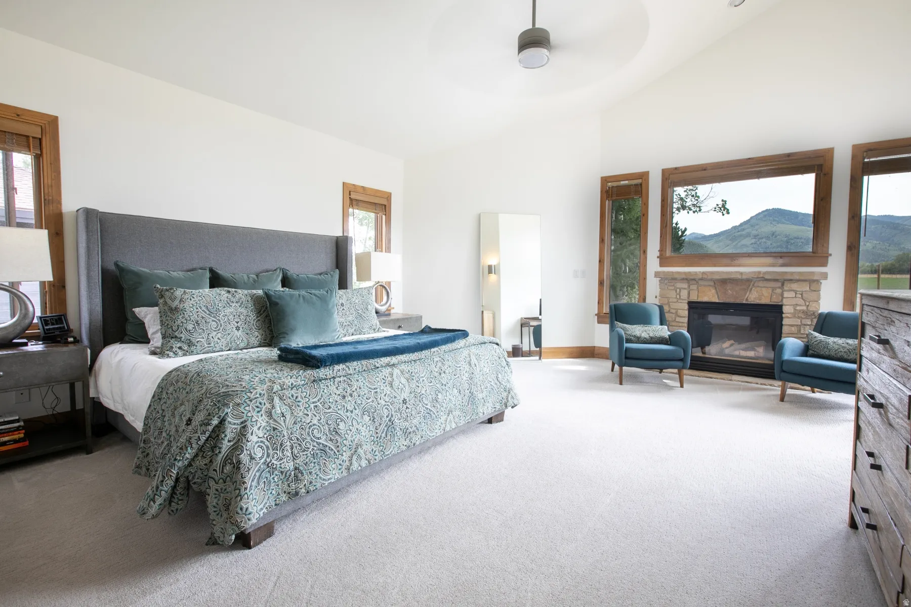 Spacious room featuring vaulted ceilings, a stone fireplace, large windows with wood trim, wall-to-wall carpeting, and a ceiling fan