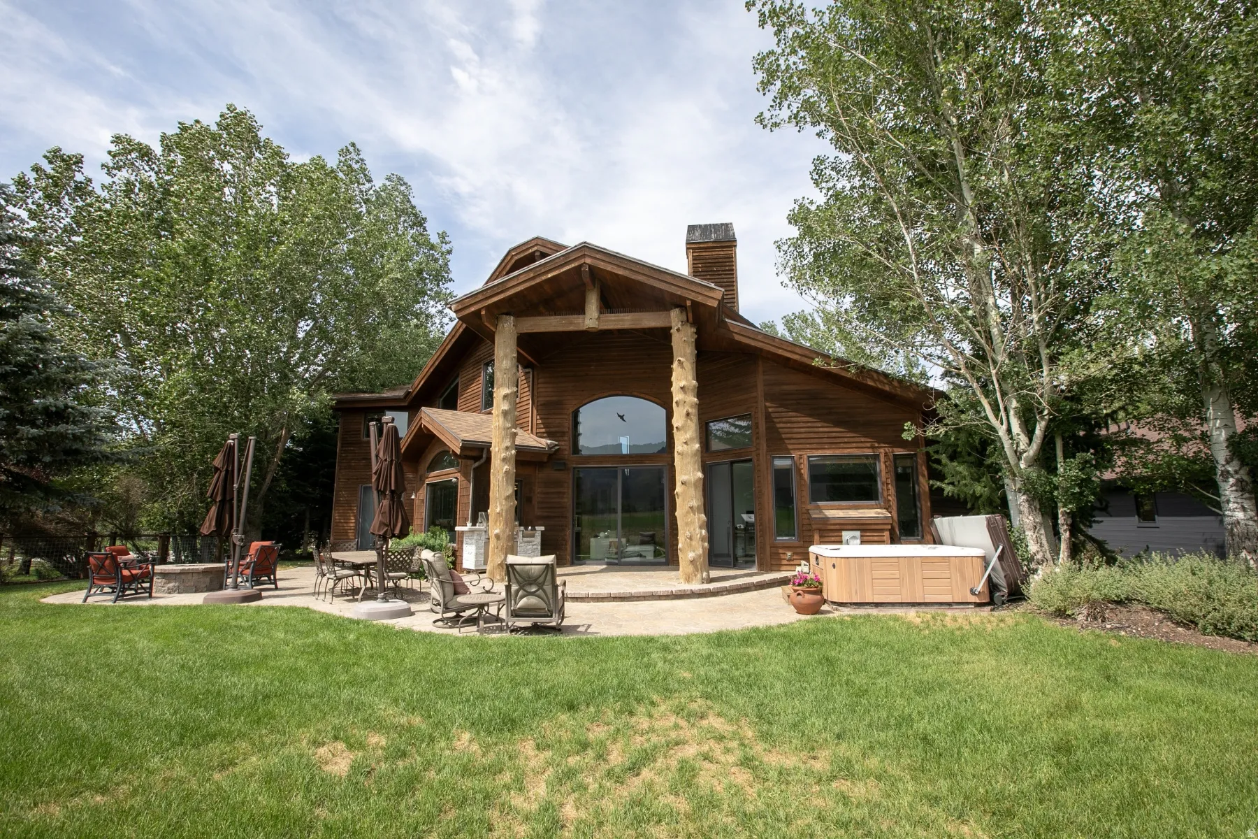 Wood-sided exterior featuring expansive windows, a large patio with a built-in fire pit, and a hot tub