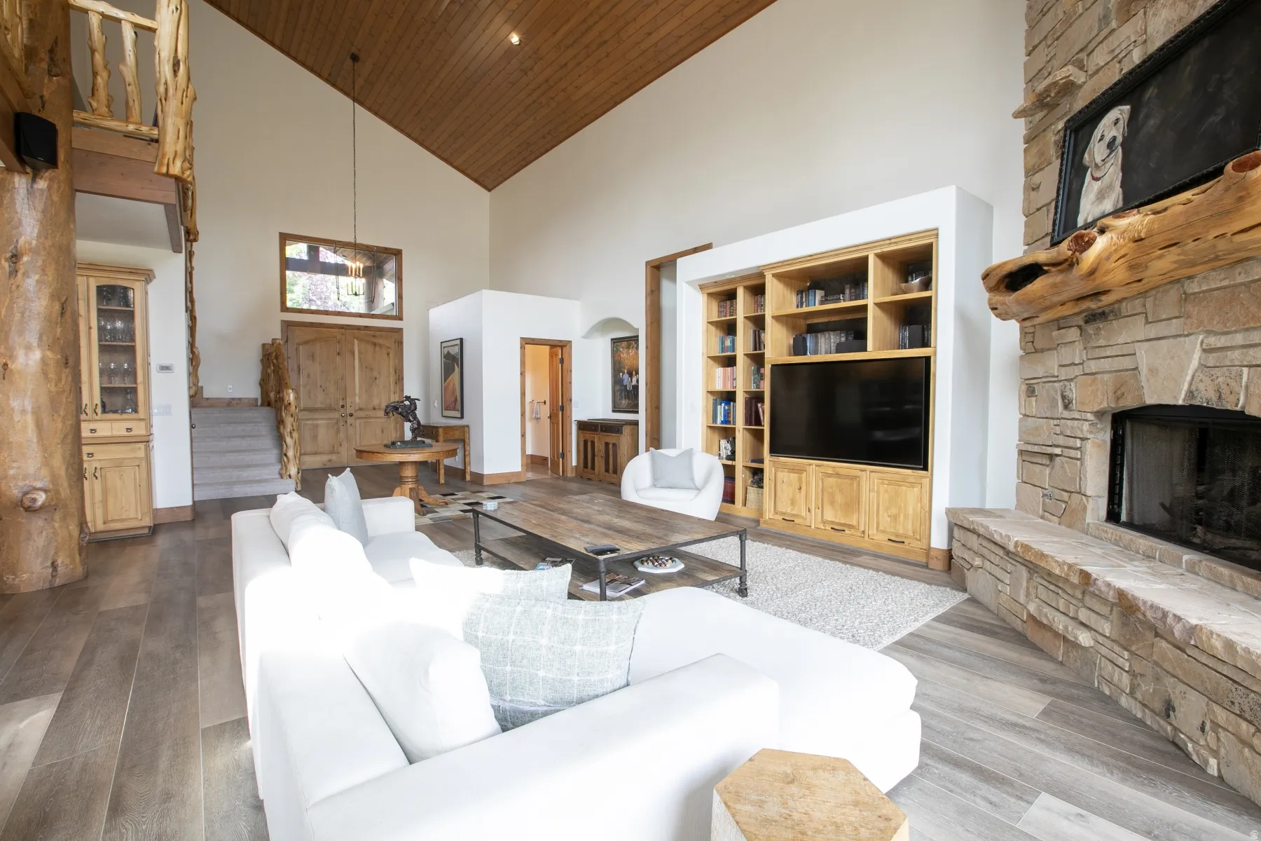 Great room featuring vaulted wood-paneled ceilings, wood-finish flooring, a stone fireplace with a substantial log mantel, a built-in entertainment center with shelving and cabinetry, and a prominent log structural column