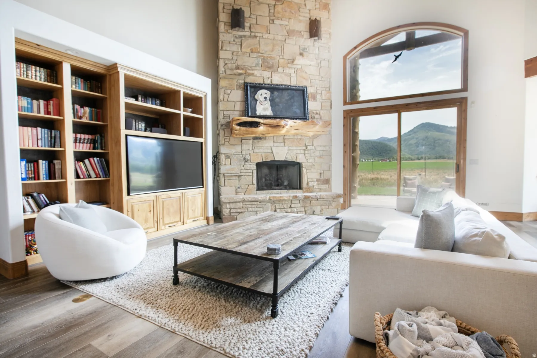 Spacious living area featuring a floor-to-ceiling stone fireplace, built-in shelving with integrated media center, wood-finish flooring, and large sliding glass doors with an arched transom window opening to a mountain landscape