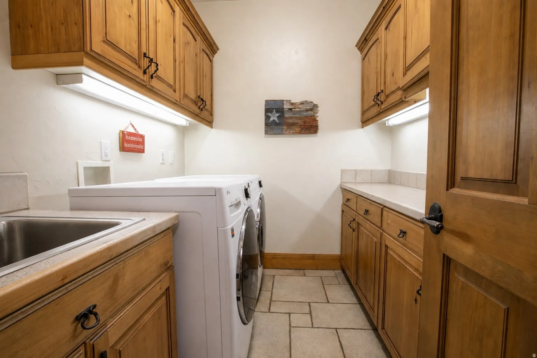 Laundry area featuring light wood cabinetry, a stainless steel sink, light-colored countertops, tiled flooring, and under-cabinet lighting