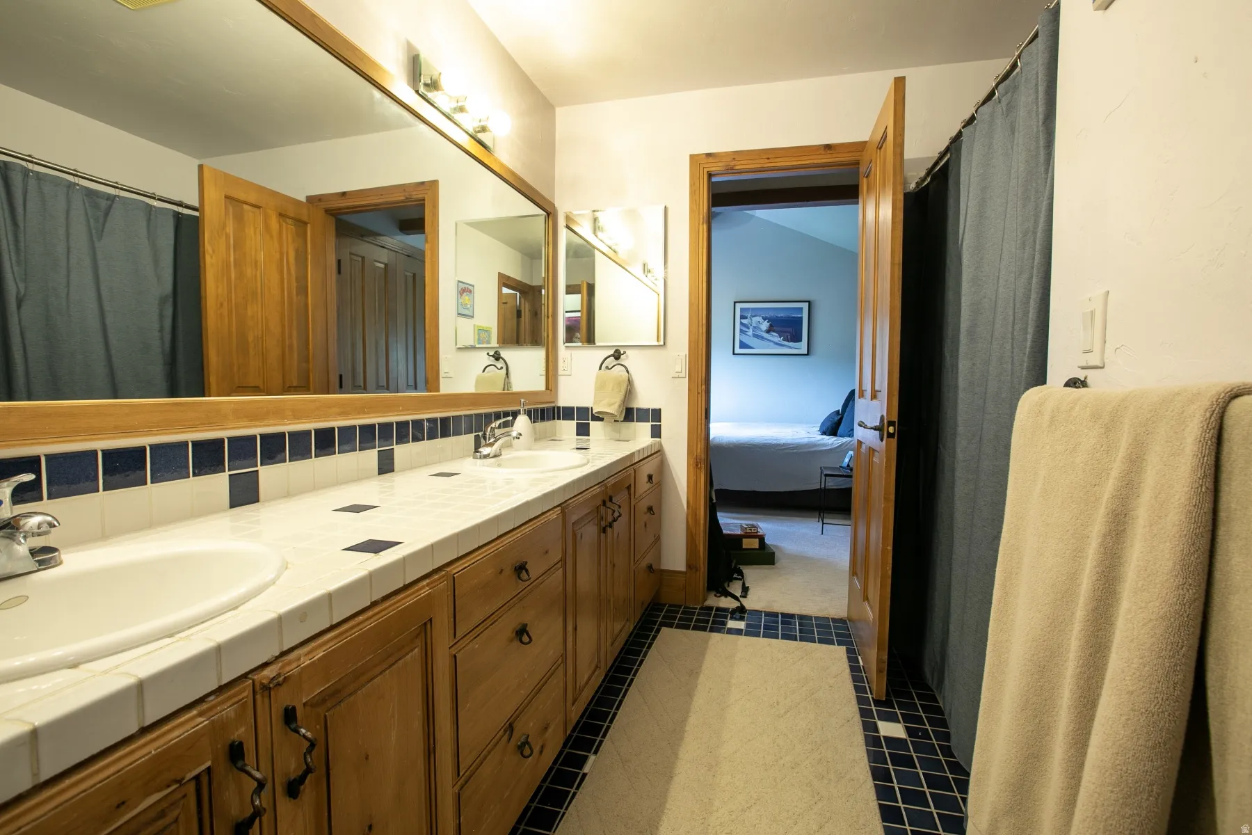 Bathroom featuring a wood-finish double vanity with dual sinks, a tile countertop with blue accent tiles, and a large framed mirror