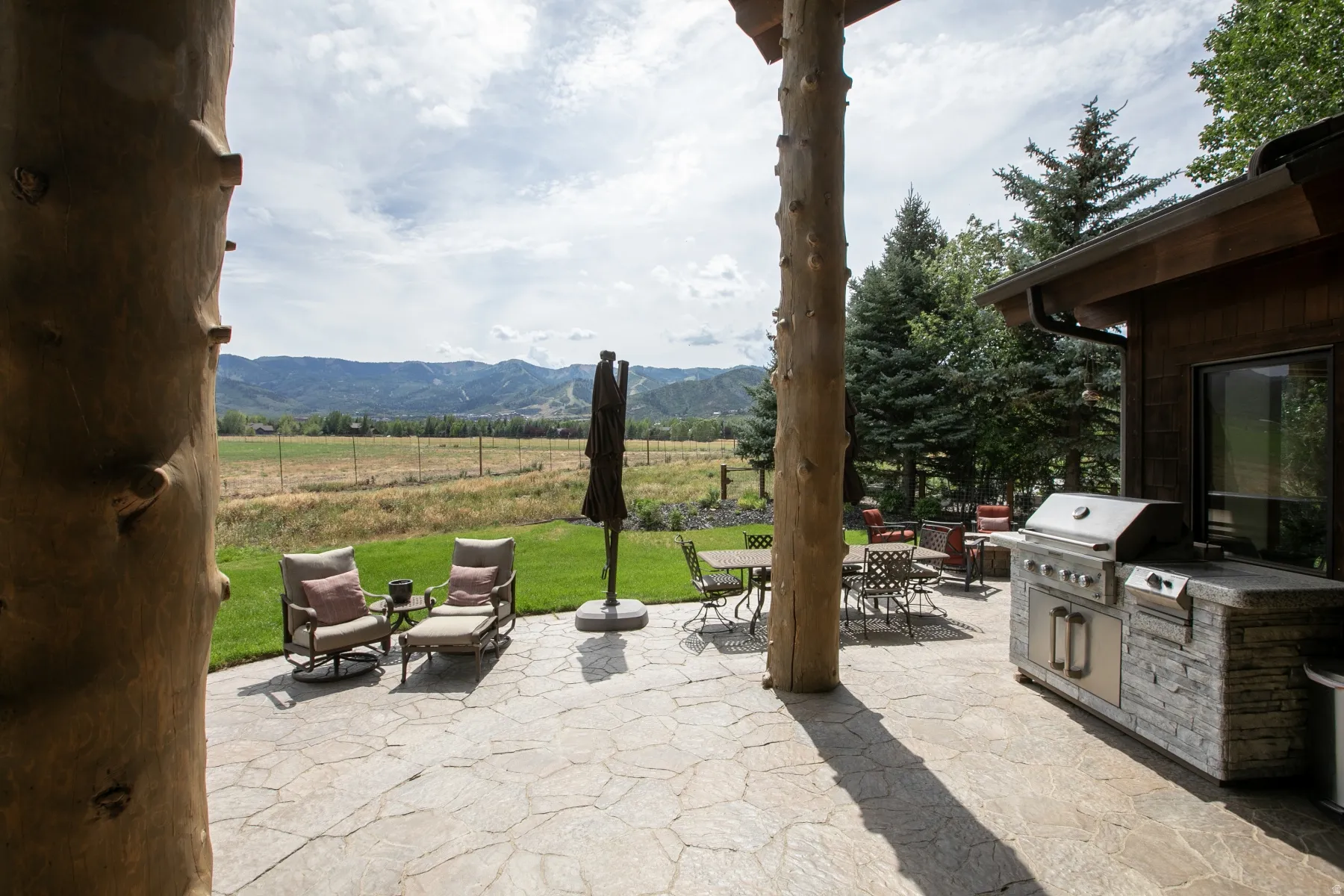 Spacious outdoor patio featuring flagstone paving, a built-in BBQ island with a stainless steel grill, and exposed timber columns supporting an overhang