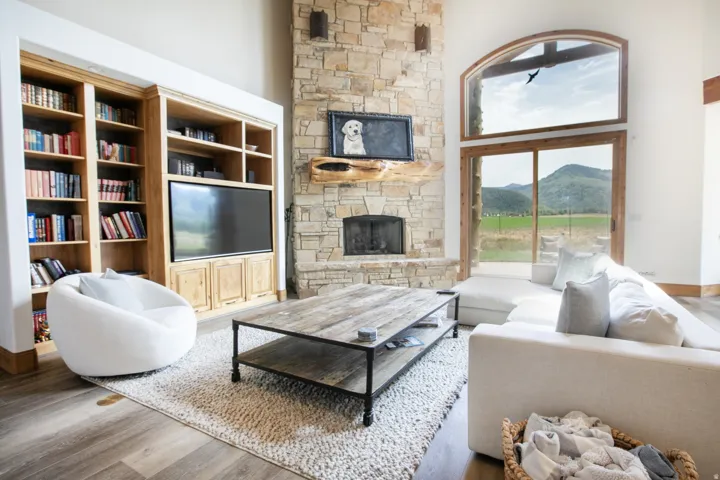 Spacious living area featuring a floor-to-ceiling stone fireplace, built-in shelving with integrated media center, wood-finish flooring, and large sliding glass doors with an arched transom window opening to a mountain landscape