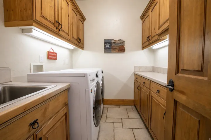 Laundry area featuring light wood cabinetry, a stainless steel sink, light-colored countertops, tiled flooring, and under-cabinet lighting