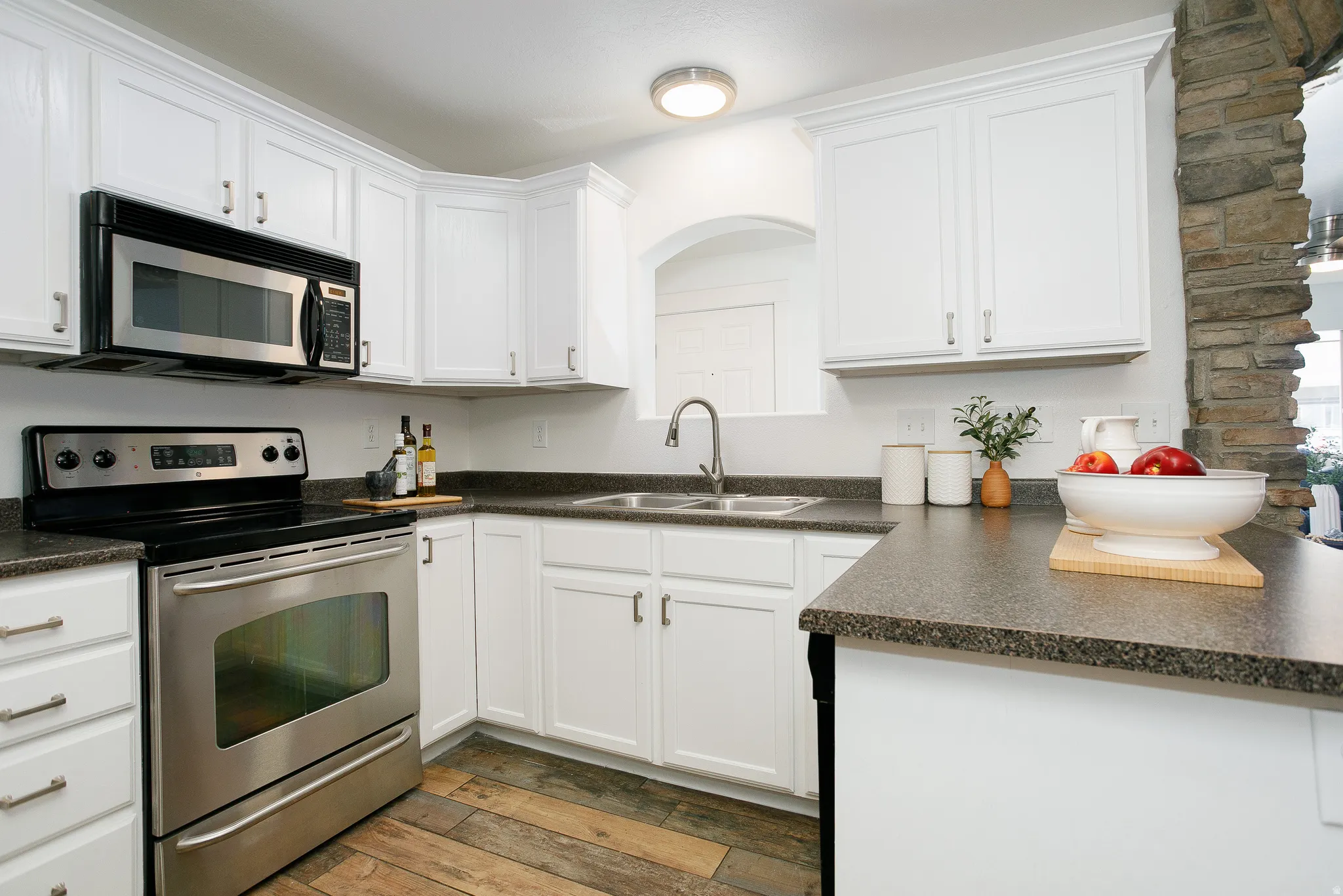 Kitchen featuring white cabinetry, stainless steel appliances, dark countertops, wood-finish flooring, and a stone accent column