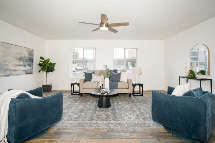 Spacious living area featuring neutral walls, wood-finish flooring, a ceiling fan with integrated lighting, and dual windows providing natural light