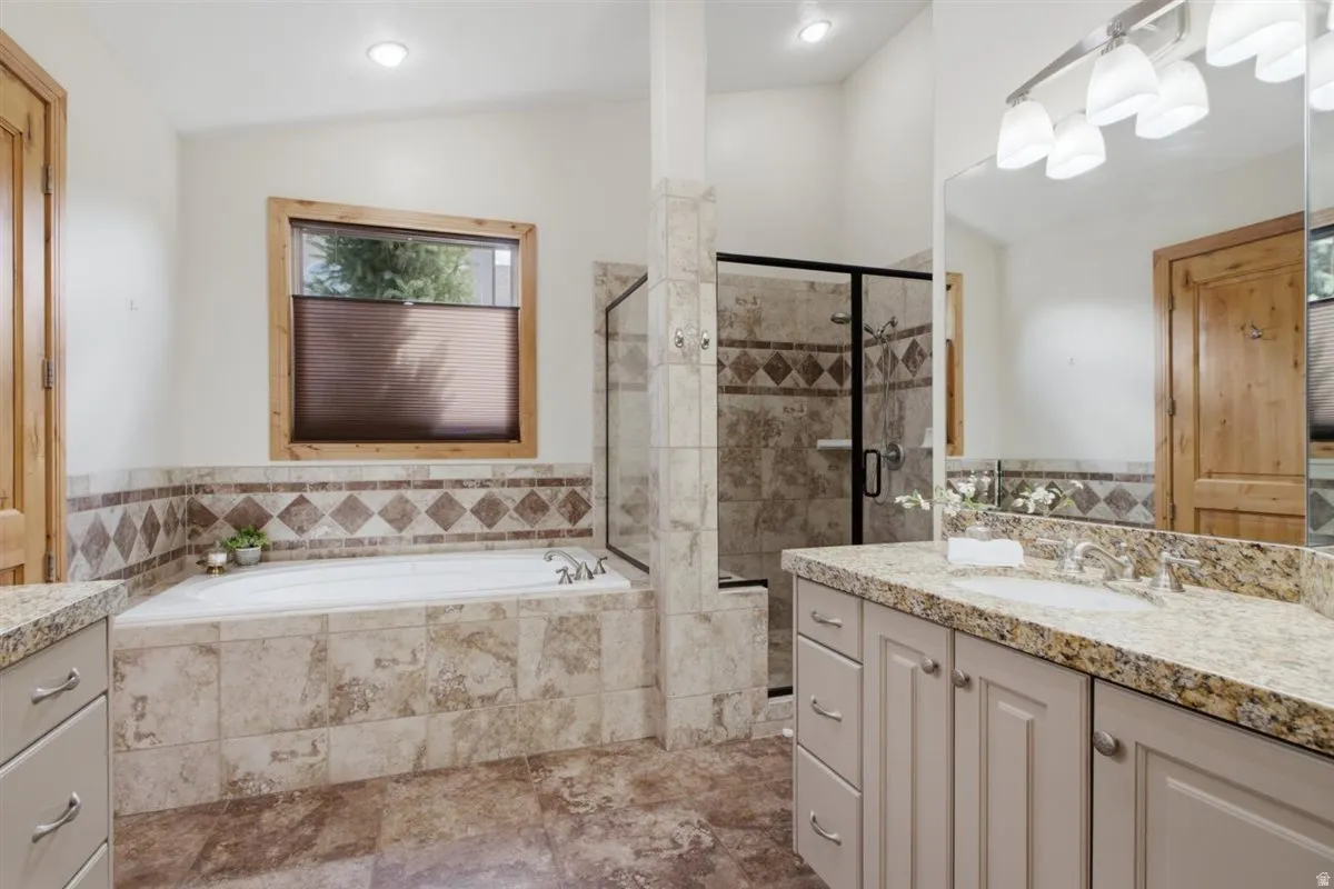 Bathroom featuring a built-in tub with tile surround, a separate glass-enclosed shower, a double vanity with stone countertops, and wood-paneled doors