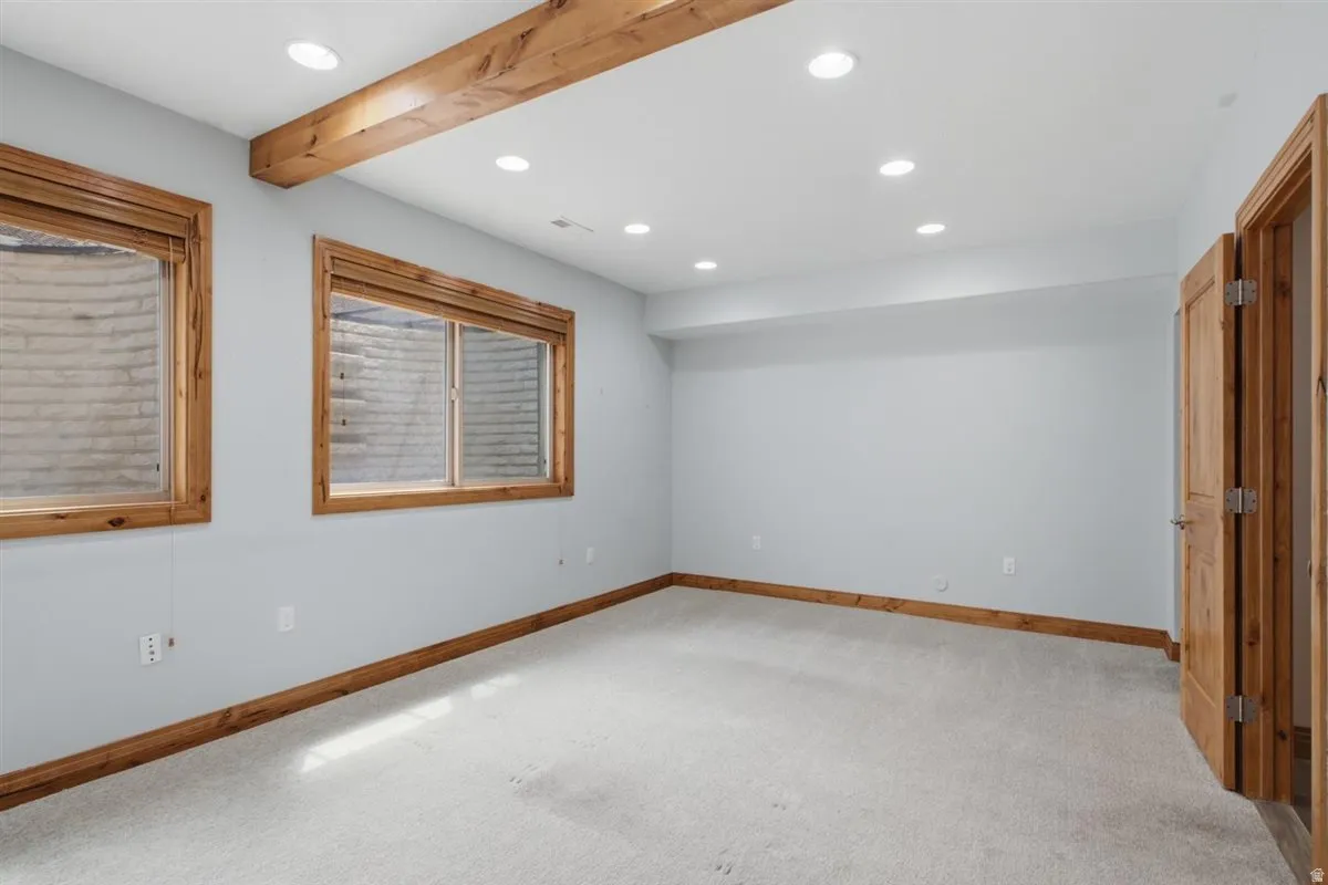 Carpeted room featuring two egress windows with wood trim, a wood-finish ceiling beam, recessed lighting, and wood-paneled doors