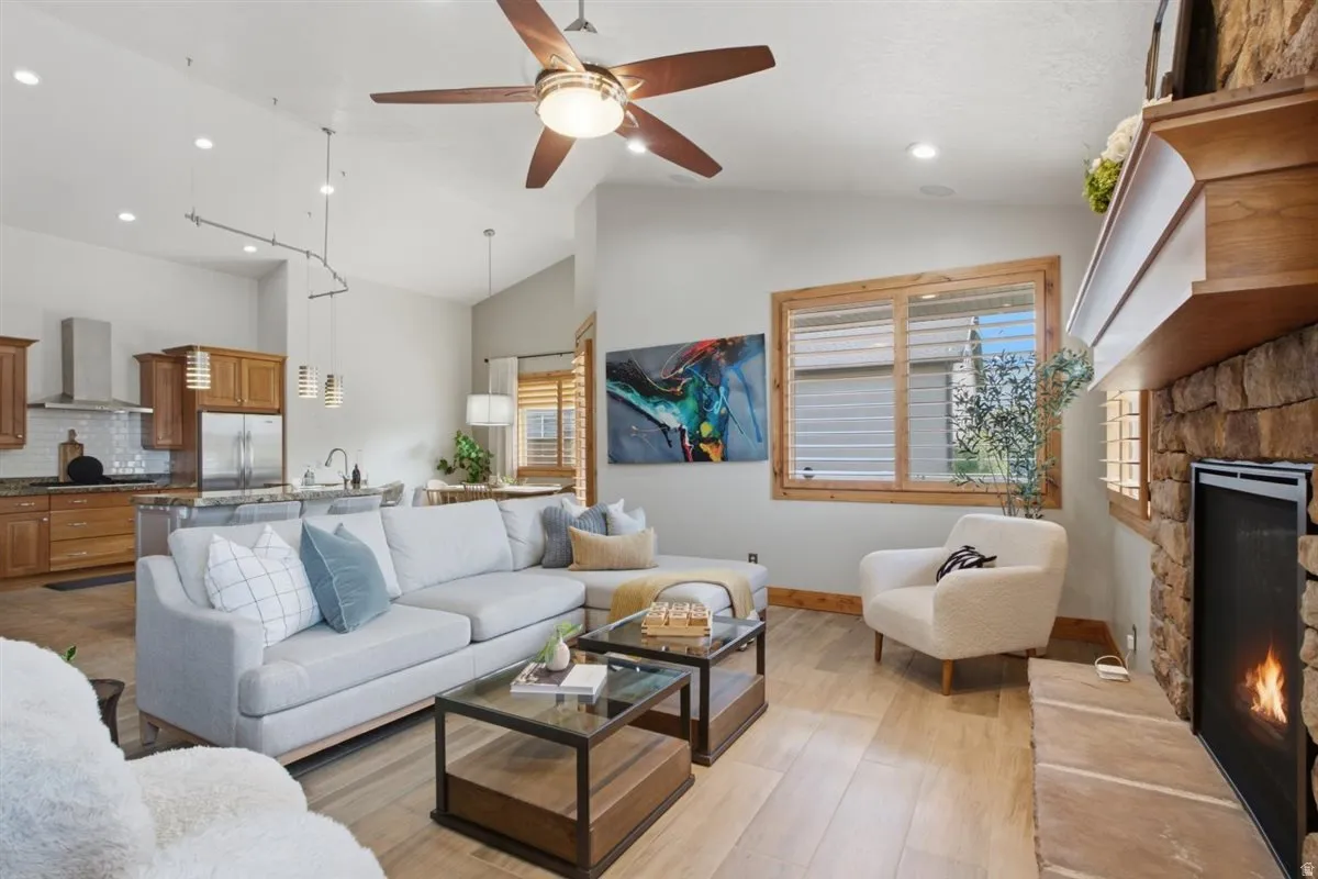 Vaulted ceiling living area with a stone fireplace, wood-finish flooring, and a ceiling fan