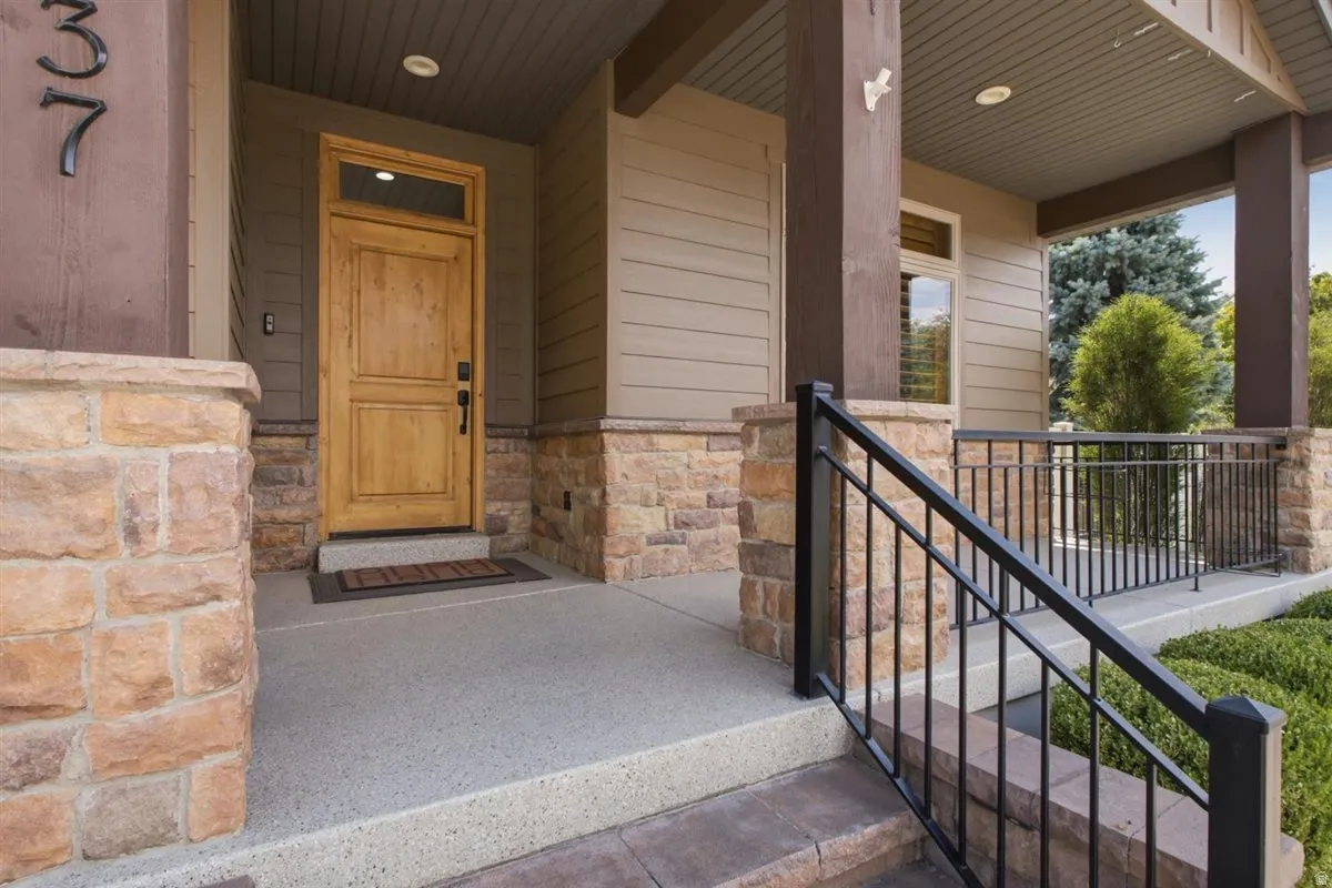 Welcoming front entry featuring a natural wood door with transom window, stone veneer accents, durable concrete porch, wrought iron railing, and recessed lighting