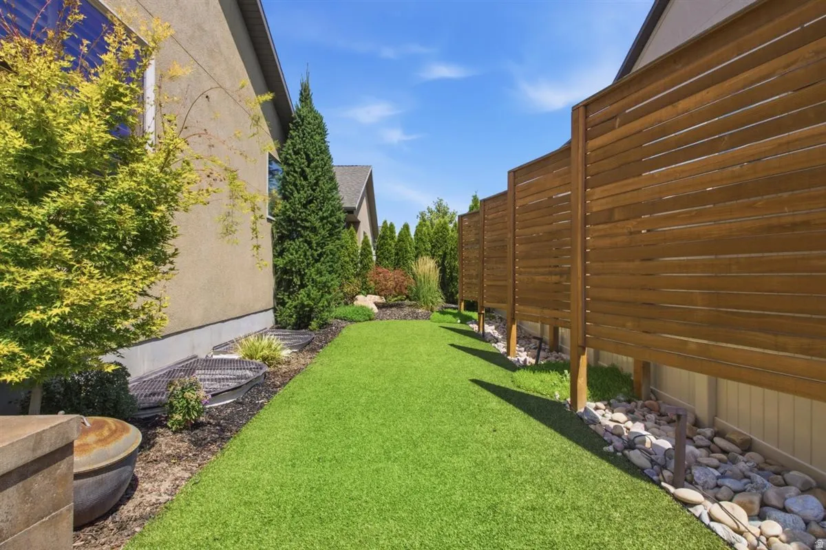 Landscaped side yard featuring artificial turf, a horizontal wood fence, mature trees, and a rock bed