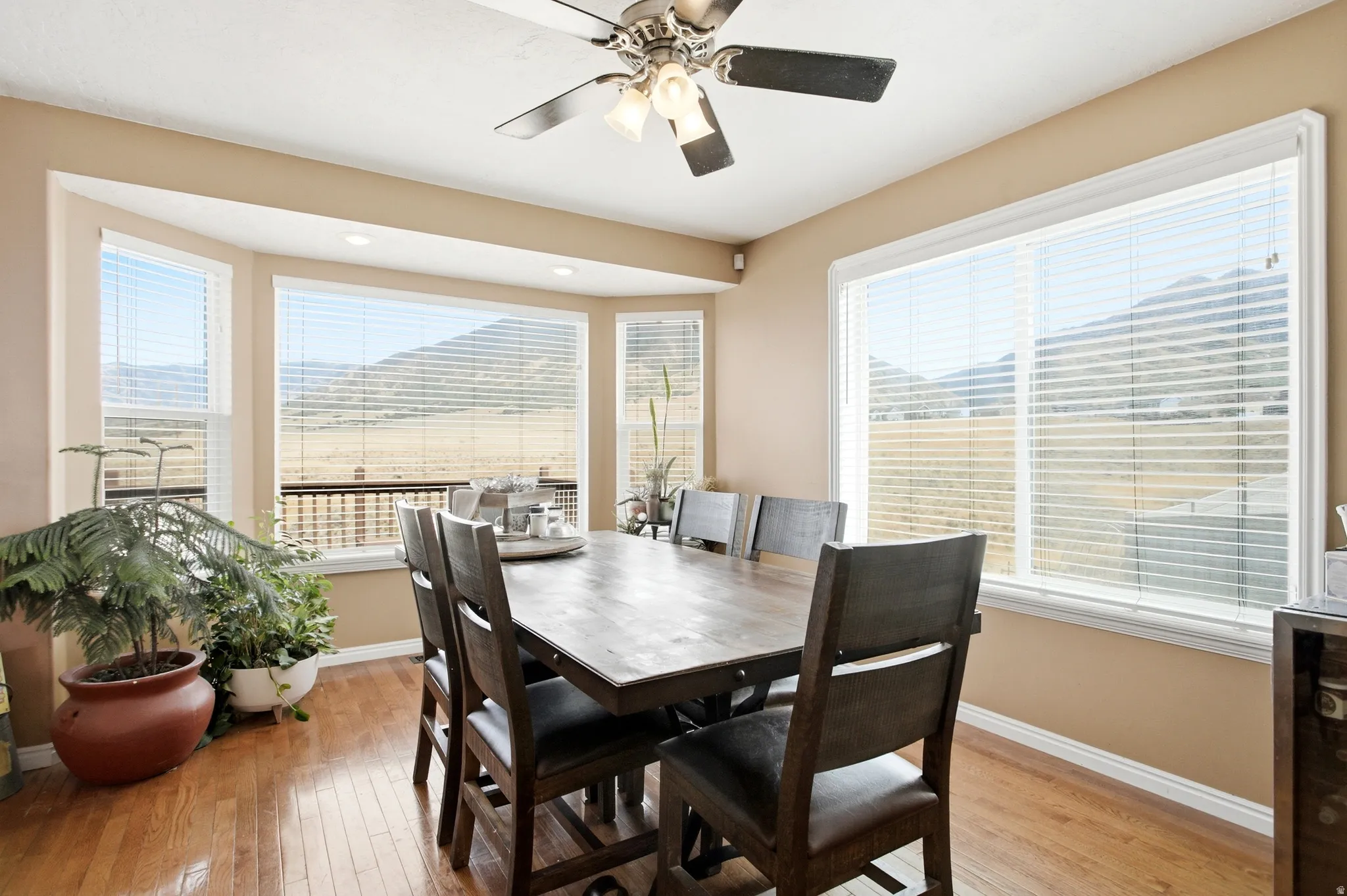 Bright dining room featuring expansive windows with cellular shades, hardwood flooring, ceiling fan with integrated lighting, and ample natural light overlooking the surrounding landscape